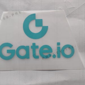 May include: A white sticker with the teal text "Gate.io" and a teal logo. The logo is a stylized letter "G" with a square inside. The sticker is on a white surface.