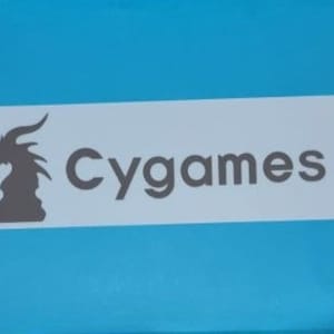 May include: A white rectangular sticker with a black silhouette of a dragon's head on the left and the text "Cygames" in black on the right.