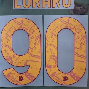 Peut inclure: Un numéro de maillot de football jaune et rouge avec le nom "LUKAKU" et le numéro "90" imprimé dessus.