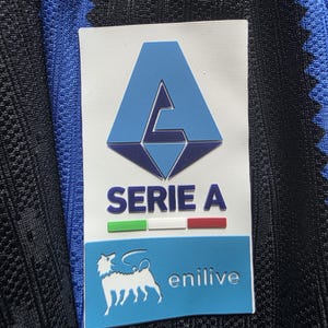 May include: A rectangular Serie A patch on a black and blue textured fabric. The patch features a light blue A logo, the text SERIE A, a green, white, and red horizontal bar, and a blue section with a white dog silhouette and the text enilive.