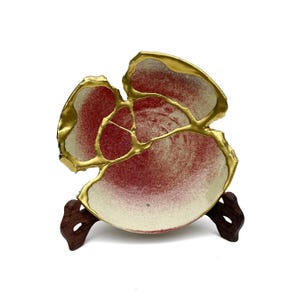 May include: A red and white ceramic plate with gold accents. The plate has an irregular shape and is displayed on a wooden stand.