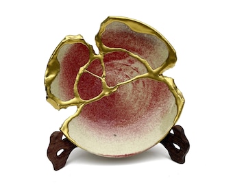 May include: A circular ceramic bowl featuring a speckled red and off-white interior surface. The bowl is broken into several segments that are rejoined with thick, raised gold-colored lines. It is displayed on a dark brown wooden stand.