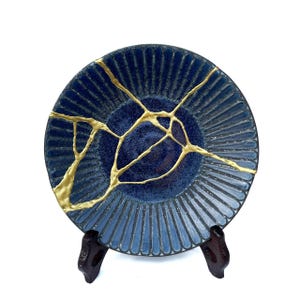 May include: A blue ceramic plate with gold-colored kintsugi repair lines. The plate has a textured, ridged design and is displayed on a dark wooden stand. The center of the plate is a darker shade of blue.