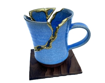 Peut inclure: Une tasse en céramique bleu clair avec une finition émaillée craquelée et une ligne de réparation dorée courant verticalement le long du côté et du bord. La tasse présente une anse incurvée et une base brun foncé sur un sous-verre carré en bois.