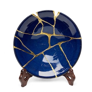 May include: A deep blue ceramic bowl featuring a kintsugi repair technique with prominent gold-colored veins filling the cracks across the surface. The bowl is displayed on a dark brown wooden stand, showcasing the contrast between the blue and gold.
