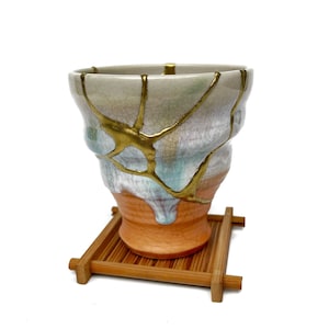 May include: A ceramic cup featuring a kintsugi design with gold-coloured lacquer filling cracks across a multi-coloured surface of cream, light blue, and orange. The cup rests on a small, square, slatted wooden stand with a natural brown finish.