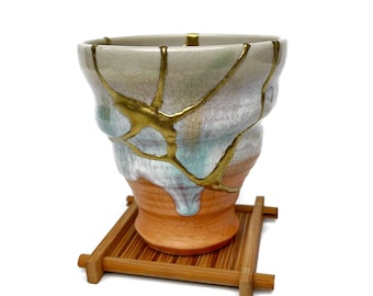 May include: A ceramic cup featuring a kintsugi design with gold-colored lacquer filling cracks across a multi-colored surface of cream, light blue, and orange. The cup rests on a small, square, slatted wooden stand with a natural brown finish.