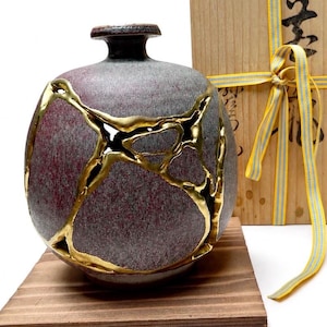 May include: A round ceramic vase with a textured, mottled purple and gray surface, featuring prominent gold-colored kintsugi repair lines. The vase sits on a wooden base next to a light-colored wooden box tied with a yellow and blue striped ribbon.