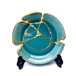 May include: A teal ceramic plate with gold-colored kintsugi repair work. The plate is displayed on a black stand, showcasing the traditional Japanese art of repairing broken pottery with gold.