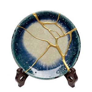 May include: A round ceramic plate featuring a gradient of deep blue to cream white, repaired with prominent gold-colored lacquer lines in a branching pattern. The plate is displayed upright on a dark brown wooden stand.