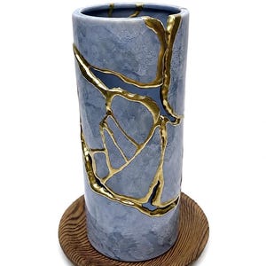 May include: A cylindrical ceramic vase with a mottled light blue finish, featuring prominent gold-colored cracks that run vertically along the exterior surface. The vase is positioned upright on a circular wooden base with a visible grain pattern.