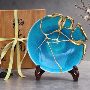 May include: A bright turquoise bowl featuring gold-colored cracks across its surface, displayed on a dark wooden stand. Beside the bowl sits a light brown wooden gift box tied with a yellow and blue striped ribbon, featuring black Japanese text.
