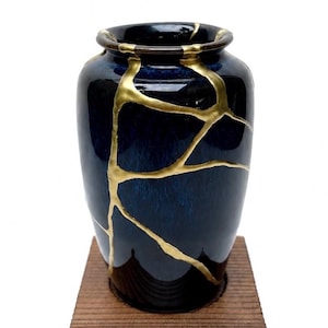 May include: A deep blue ceramic vase featuring a glossy finish and prominent gold-colored veins that trace across the surface, highlighting a repaired aesthetic. The vase has a rounded body and a short, thick neck with a smooth, circular rim.