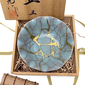 May include: A light blue ceramic bowl featuring a crackle glaze finish and gold-colored repair lines across the surface. The bowl is nestled inside a square wooden box filled with shredded paper packing material, with a wooden lid nearby.