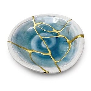 Op de afbeelding: Een ronde keramische schaal met een blauw-witte craquelé glazuurafwerking. Het oppervlak is gesegmenteerd door opvallende goudkleurige lijnen die het pad van de breuken over de kom volgen, wat een patroon van onderling verbonden aders creëert.