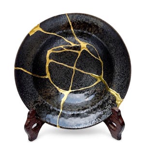 May include: A black ceramic plate with gold-colored kintsugi repair lines. The plate is displayed on a dark wooden stand. The kintsugi technique highlights the history of the plate, making it a unique decorative piece.