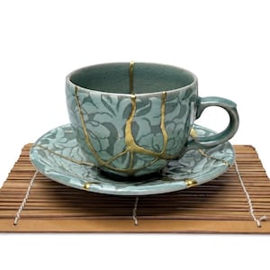 May include: A light blue-green ceramic teacup and saucer set featuring a floral pattern and gold-coloured cracks across the surface. The cup has a curved handle and a matching saucer, both decorated with gold lines that highlight the repaired fractures.