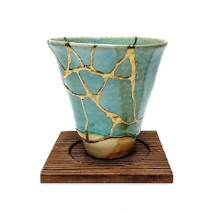 Puede incluir: Una taza de cerámica cónica con una superficie esmaltada en turquesa y azul claro, con una red de grietas de color dorado por todo el exterior. La taza tiene una base cónica y descansa sobre un posavasos cuadrado de madera marrón oscuro.