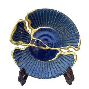 May include: A blue ceramic plate with a gold-colored kintsugi repair. The plate has a fan-like pattern and is displayed on a dark wooden stand. The plate is approximately 8 inches in diameter.