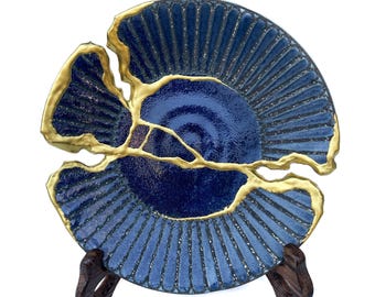 May include: A circular dark blue ceramic plate featuring a textured, ribbed surface and a glossy finish. The plate is broken into two distinct pieces that are joined together by thick, raised gold-coloured lines along the fracture seams.
