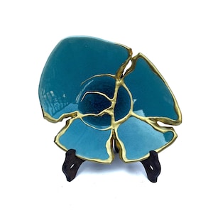 May include: A teal blue ceramic bowl with gold accents, resembling a flower, on a wooden stand.