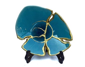 May include: A turquoise ceramic bowl featuring a cracked design with gold-coloured seams connecting the broken pieces. The bowl has a glossy finish with a visible crackle pattern in the centre and is displayed on a dark brown wooden stand.