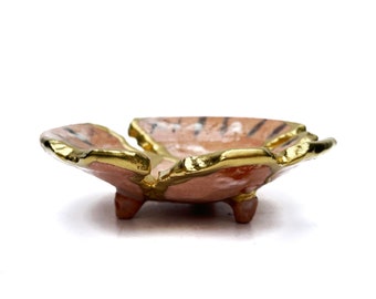 May include: A small, shallow ceramic bowl with a flower-like shape, featuring a textured, light pink interior with dark streaks and a metallic gold-painted rim. The bowl is supported by three short, stubby feet and has a glossy finish.