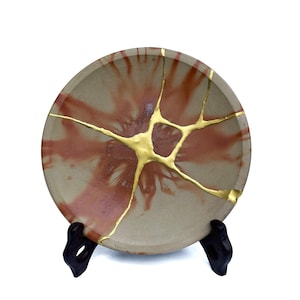 May include: A round ceramic plate with a beige and reddish-brown design, featuring gold-colored kintsugi repairs. The plate is displayed on a black stand, showcasing the traditional Japanese art of repairing broken pottery.