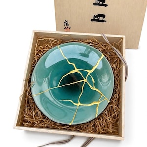 May include: A teal ceramic plate with gold-colored kintsugi repair lines, presented in a wooden box with straw and a decorative ribbon. The plate has a gradient color, transitioning from a lighter teal to a darker teal in the center.
