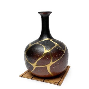 May include: A round ceramic vase with a narrow neck, featuring a dark brown and black textured glaze. The surface is decorated with prominent gold-colored lines that create a cracked pattern across the body of the vessel.