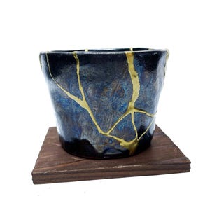 May include: A dark blue ceramic cup with gold-colored kintsugi repair lines, sitting on a small, dark wooden square. The cup has a textured surface and a unique, artistic appearance.