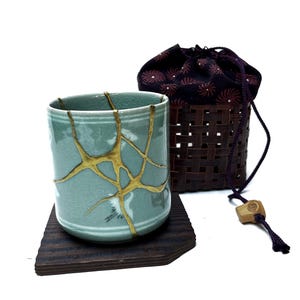 May include: A celadon ceramic cup with gold kintsugi repair lines, on a dark wooden coaster. Behind it is a woven basket with a drawstring bag. The bag is black with a floral pattern.
