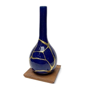 May include: A deep blue ceramic vase with a long, slender neck and a rounded base. The surface features prominent gold-coloured veins that create a cracked pattern across the body. The vase is positioned on a square, light brown wooden base.