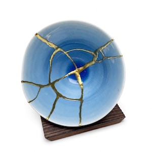 May include: A round ceramic bowl featuring a light blue gradient finish that deepens to a dark blue center. The bowl is repaired with gold-colored lacquer lines in a kintsugi style, creating a webbed pattern across the surface of the vessel.