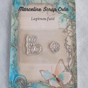 May include: A clear plastic package with a white background and a decorative border. The package contains two clear rubber stamps, one of a rabbit and one of a mushroom. The text "Marceline Scrap Crea" and "Lapinou futé" is printed on the package.