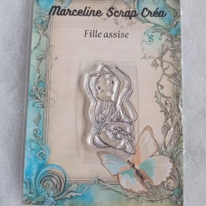May include: Clear stamp with a line drawing of a girl sitting down. The stamp is on a white card with a blue and green floral border. The text "Marceline Scrap Créa" and "Fille assise" is printed on the card.