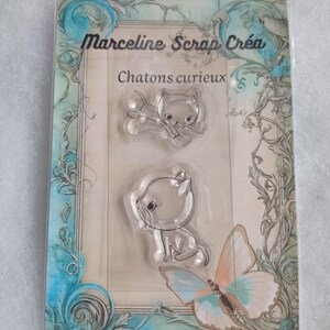 May include: A clear stamp set with two images of kittens. The set is packaged on a card with a blue and green floral design. The text "Marceline Scrap Crea" and "Chatons curieux" is printed on the card.