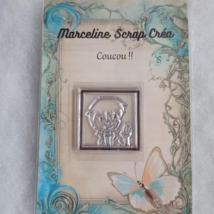 May include: A clear plastic package with a white background featuring a cartoon character in a square frame. The character is waving with a smile. The text "Marceline Scrap Créa" and "Coucou!!" are printed on the package. A butterfly is in the bottom right corner.