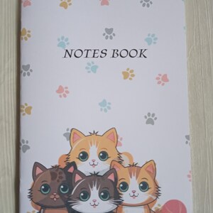 Peut inclure: Un carnet blanc avec un motif d'empreintes de pattes et le texte "NOTES BOOK" sur la couverture. La couverture présente une illustration de quatre jolis chats de dessin animé avec de la laine colorée.