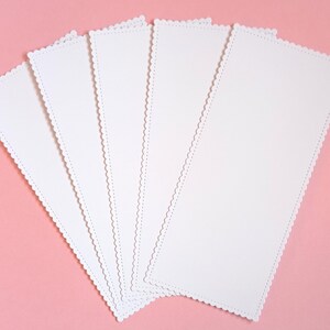 Peut inclure: Cinq marque-pages rectangulaires en papier blanc disposés en éventail. Chaque marque-page présente un bord festonné décoratif avec un détail de bordure piquée, offrant une apparence propre et uniforme pour ces articles de papeterie vierges.