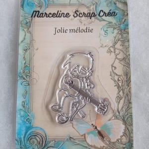 May include: Clear stamp featuring a line drawing of a person playing a guitar with the text "Jolie mélodie" above the image. The stamp is on a white background with a blue and green floral border.