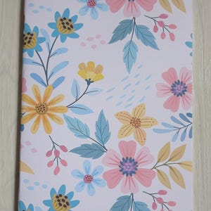 Peut inclure: Un cahier à motifs floraux sur fond blanc. Le motif présente des fleurs roses, jaunes et bleues, avec des feuilles vertes et beiges. Les fleurs sont stylisées et disposées de manière éparse sur la couverture du cahier.