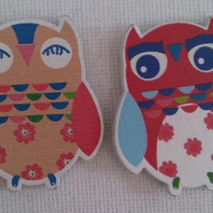 Peut inclure: Deux décorations en bois en forme de hibou avec des motifs colorés. Le hibou de gauche est beige avec des ailes rouges et un motif bleu et vert sur la poitrine. Le hibou de droite est rouge avec des ailes bleues et une poitrine blanche avec des fleurs rouges.