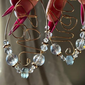 May include: Gold wire earrings with clear and light blue crystal beads. The earrings are shaped like a swirl.