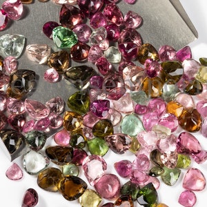 May include: A large collection of loose, faceted, pear-shaped gemstones in various shades of pink, green, brown, and clear. The gemstones are scattered on a silver tray and a white surface.