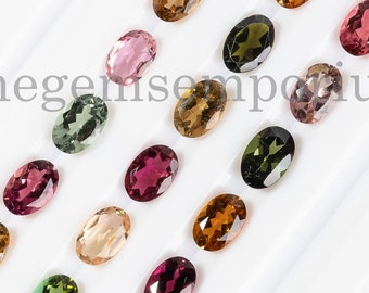 May include: Assortment of oval-shaped gemstones in various colors, including pink, green, brown, and black. The gemstones are arranged in rows on a white surface. Thegemsemporium is printed on the surface.