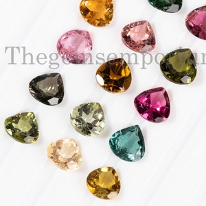 May include: Assortment of heart-shaped gemstones in various colors, including pink, green, brown, and yellow. The gemstones are arranged on a white surface.