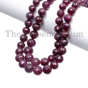 Op de afbeelding: Een streng dieprode robijn kralen, elke kraal is facetgeslepen en rond, de kralen zijn geregen op een wit koord.