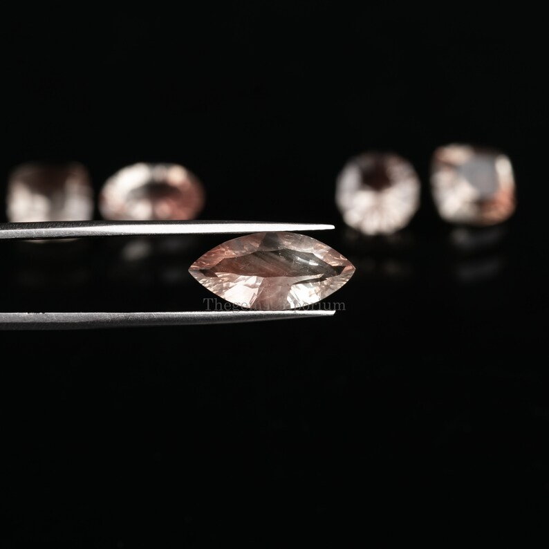 May include: A light pink marquise-shaped gemstone held in tweezers. The gemstone has a slight shimmer and is surrounded by a black background.