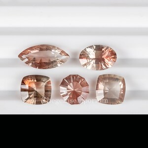 May include: Five sunstone gemstones in various shapes and sizes. The gemstones are a light orange color with a slight pink hue. The gemstones are displayed on a white surface.
